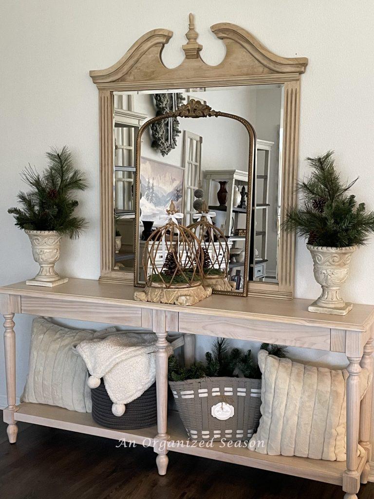 A console table with two mirrors, a cloche, and two planters with greenery sitting it on top.  Two faux fur pillows, a basket with greenery, and a basket with a throw blanket are on the bottom shelf! 