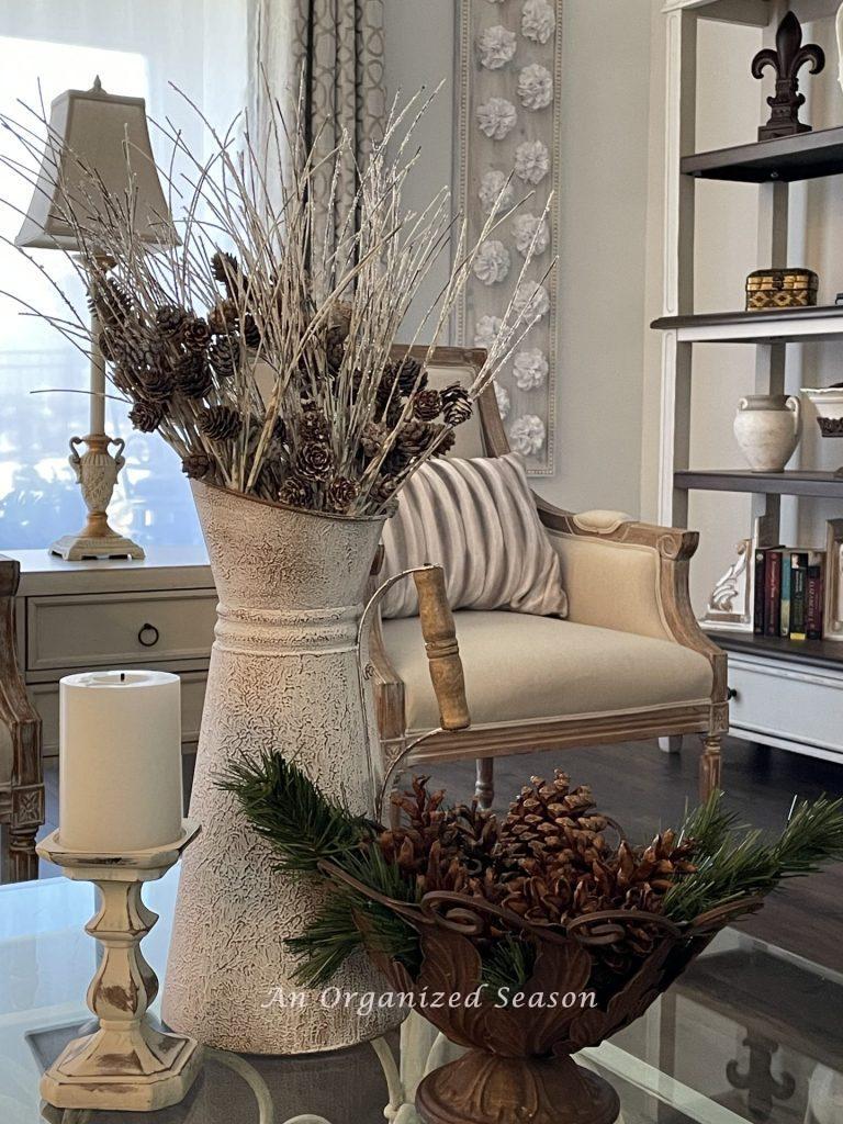 A pitcher filled with white twigs and pinecones, next to a candle, and a brown metal bowl of pinecones. 