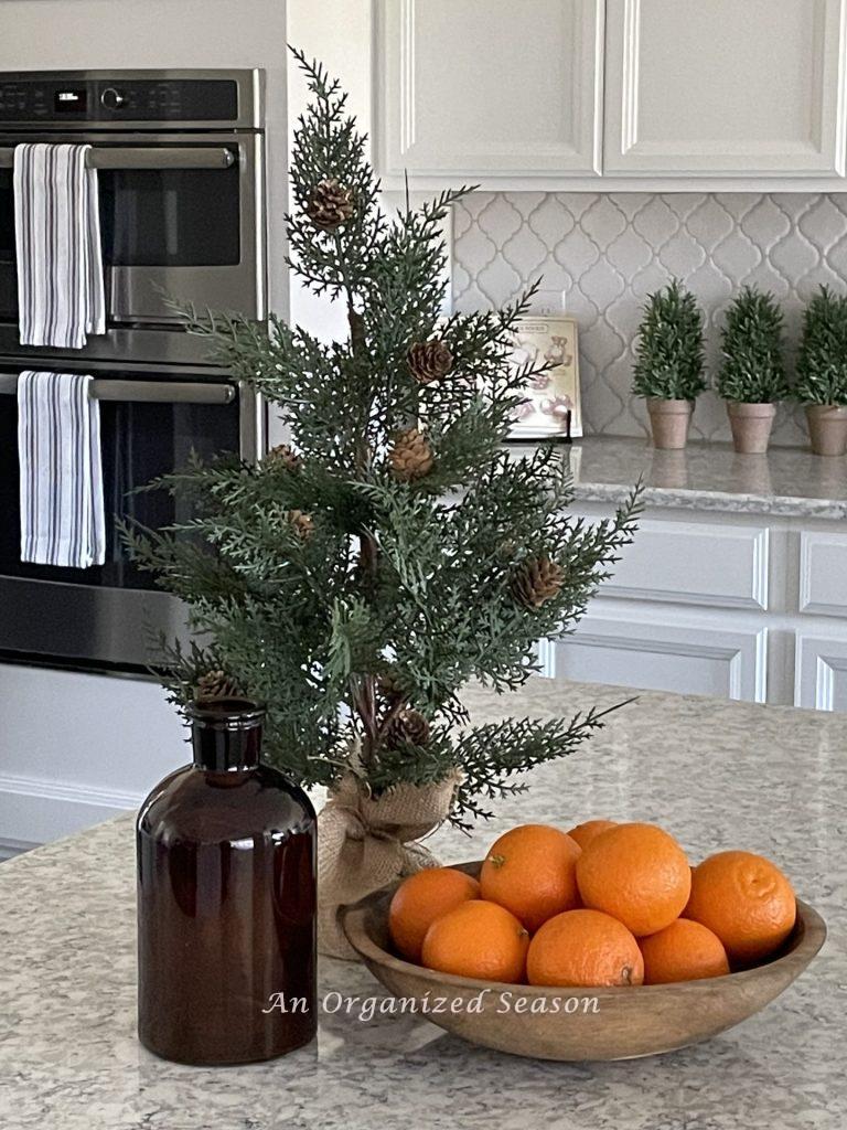 A small evergreen tree with pinecones, a brown bottle, and a wood bowl filled with oranges, on a kitchen counter. 
