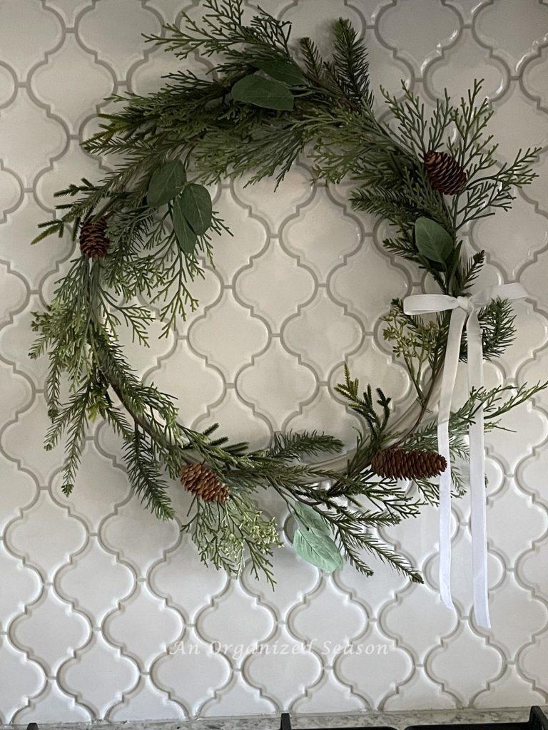 An evergreen wreath with a white velvet bow hanging on a kitchen backsplash.