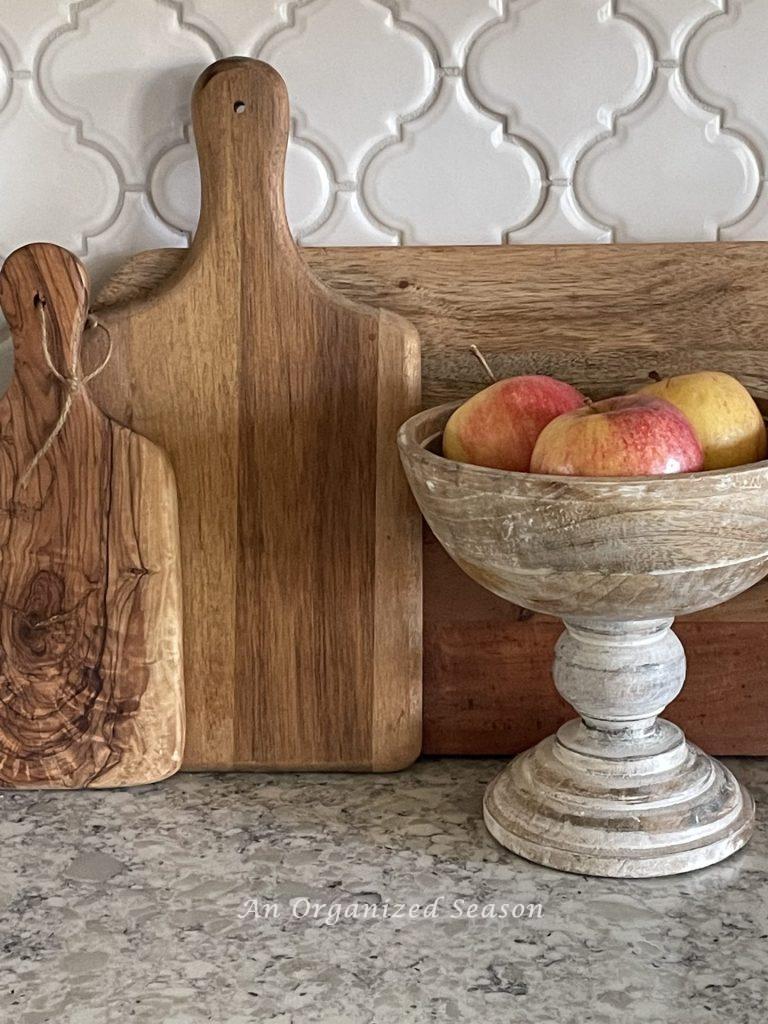 Three cutting boards stacked against the backsplash and held upright with a wooden bowl of apples allows you to declutter and decorate your kitchen counters all at once.  