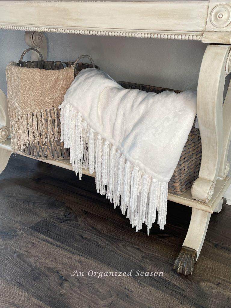 Two baskets under a table that are filled with throw blankets.