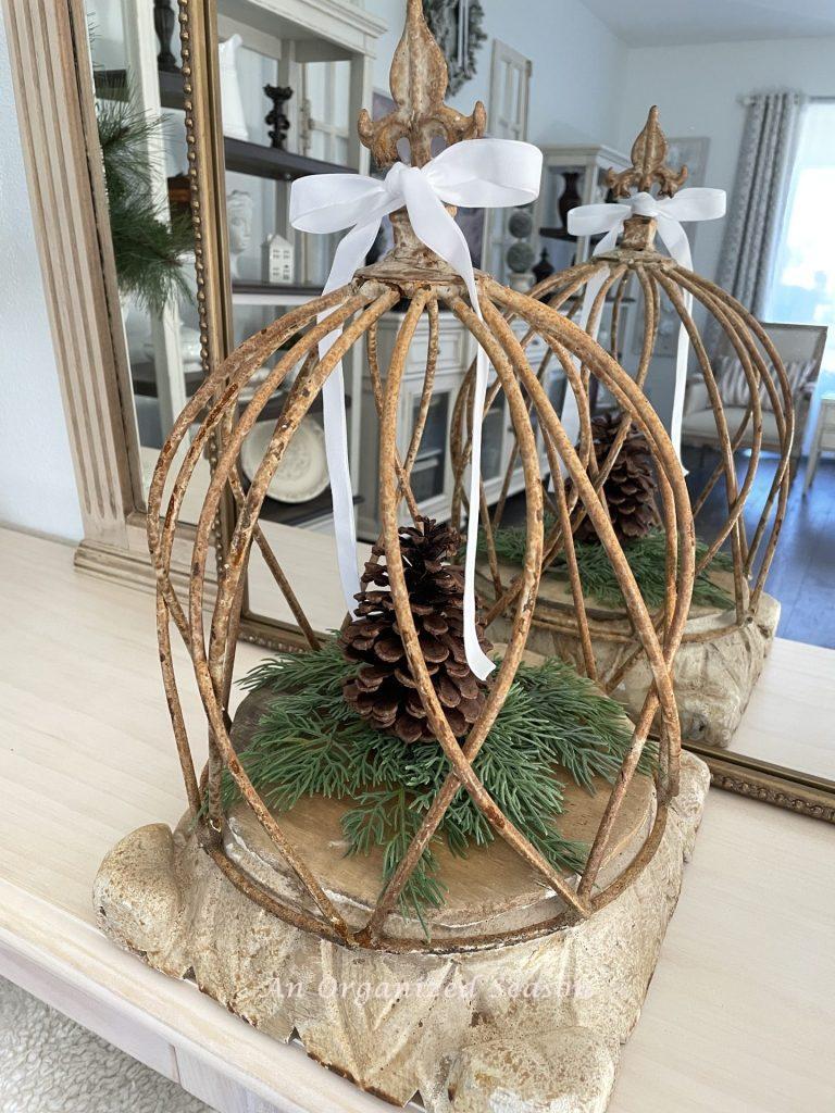 A metal cloche sitting on a wood pedestal with evergreen and a pinecone inside. 