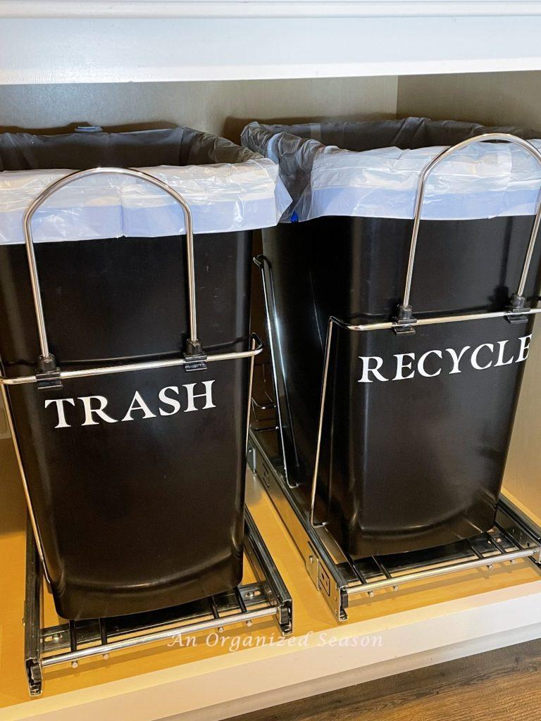Two black trash cans that pull-out from the kitchen cabinet that are labeled "trash" and "recycle."
