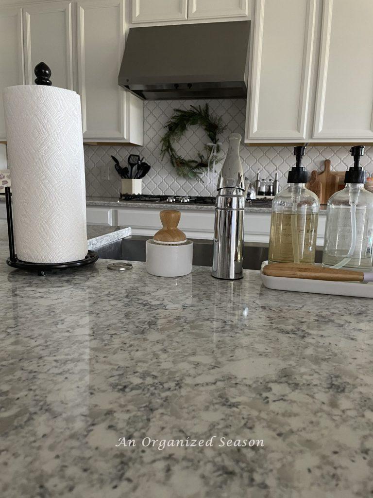 Paper towels on a decorative holder, a scrub brush, hand and dish soap sitting on a kitchen counter. 