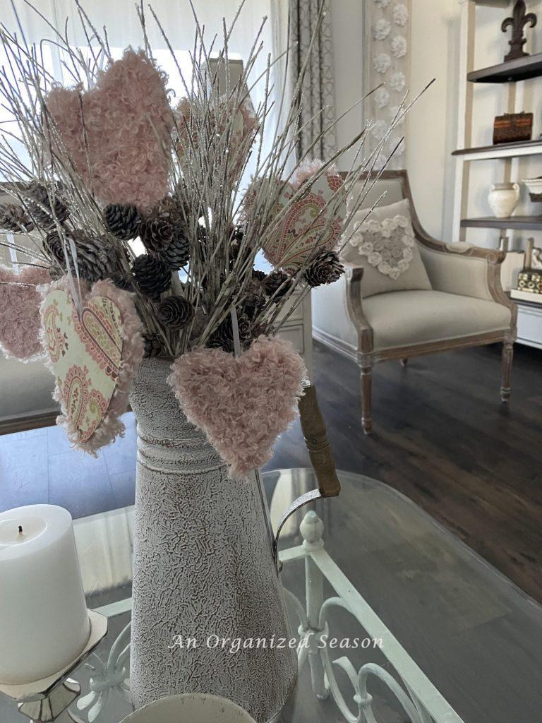 A pitcher holding white branches with pinecones that has Valentine's hearts hanging on it. 