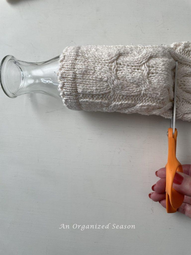 A glass vase inside the sleeve of a sweater with someone cutting off the excess with scissors, step two for how to make cozy home decor with sweaters.