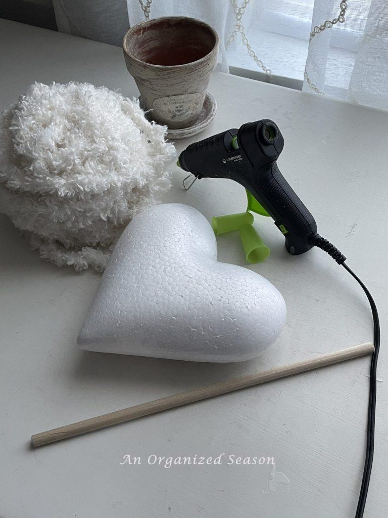 A Dollar Tree styrofoam heart, yarn, glue gun, dowel rod, and flower pot used to make a DIY topiary.
