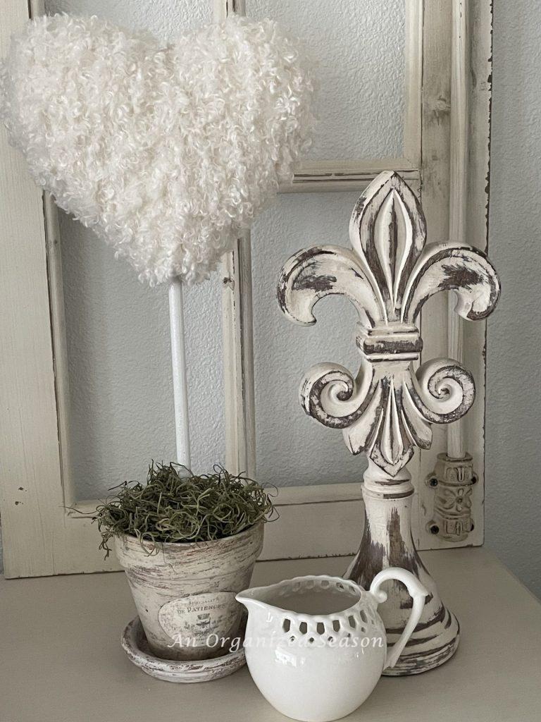 A white yarn heart shaped-topiary next to a statue and a white creamer. 