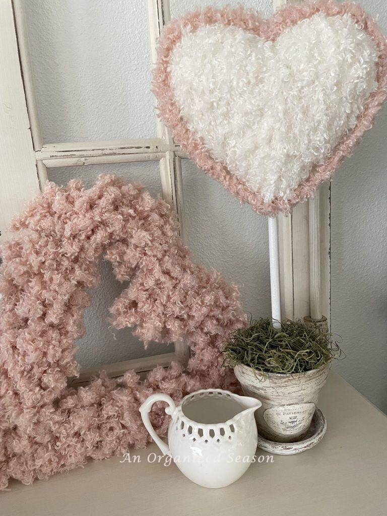 A white yarn topiary with pink edge sitting by a pink heart and a white creamer.