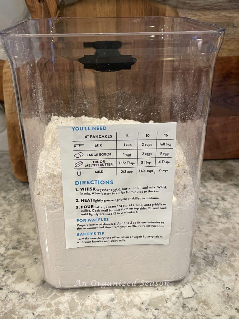 A container filled with pancake mix that has the instructions taped on the back. 