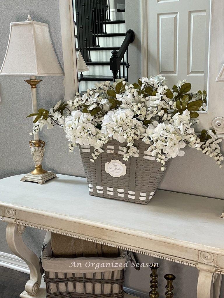 White flowers inside a gray French basket sitting on a foyer table. 