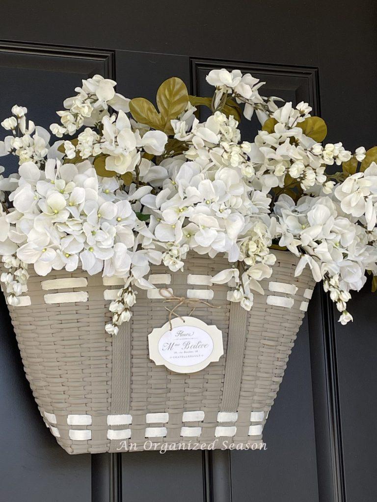 White flowers inside a gray French basket hanging on a front door. 