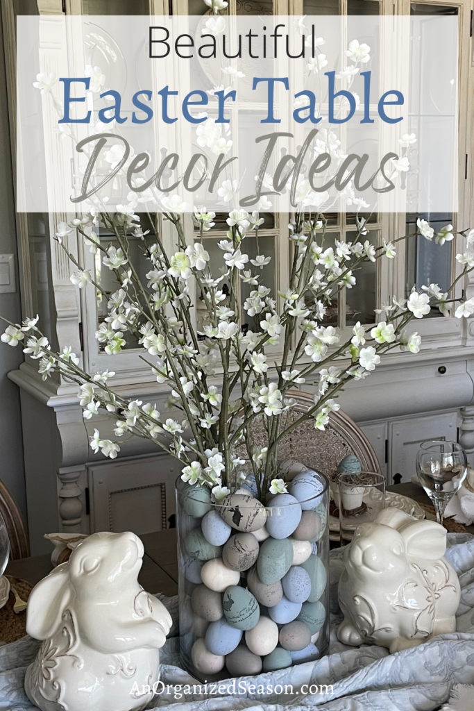 Table decorated with flowering branches in a vase filled with decorated eggs with two bunnies on either side. 