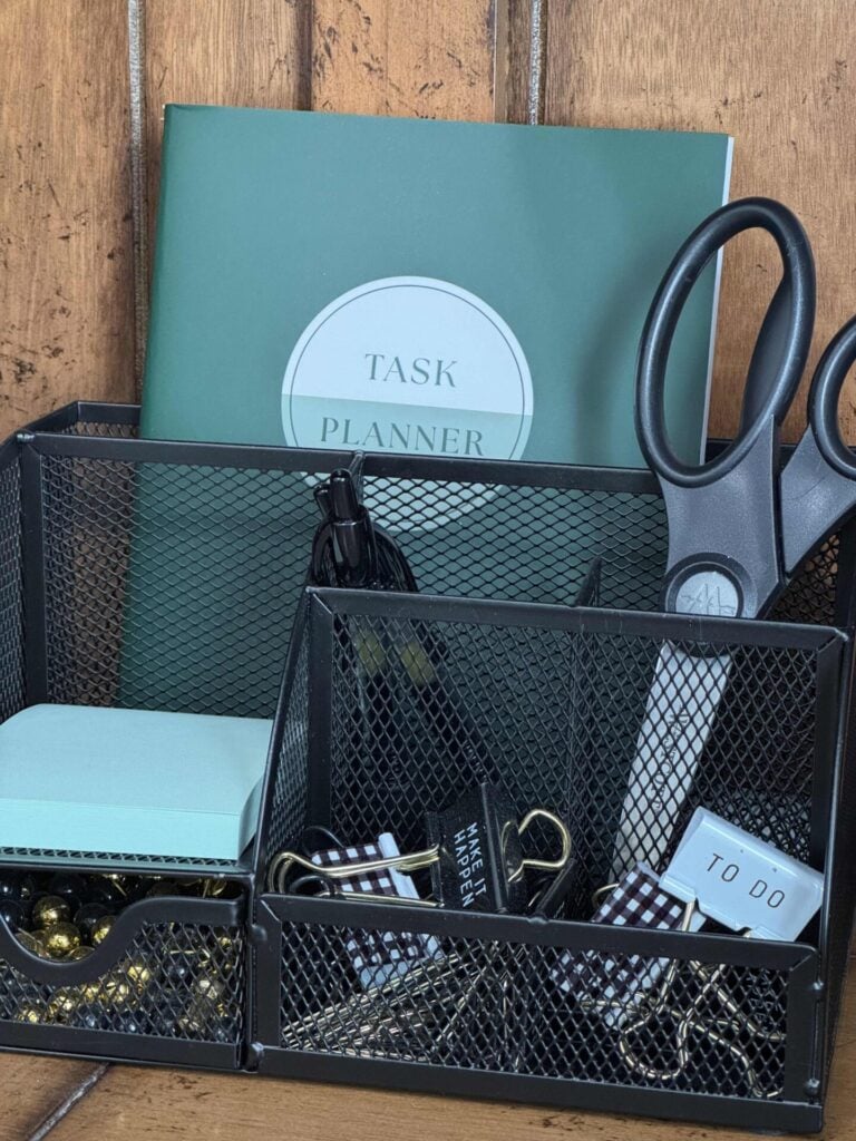 A black mesh desk organizer storing a planner, scissors, pens, post it notes, and clips.  
