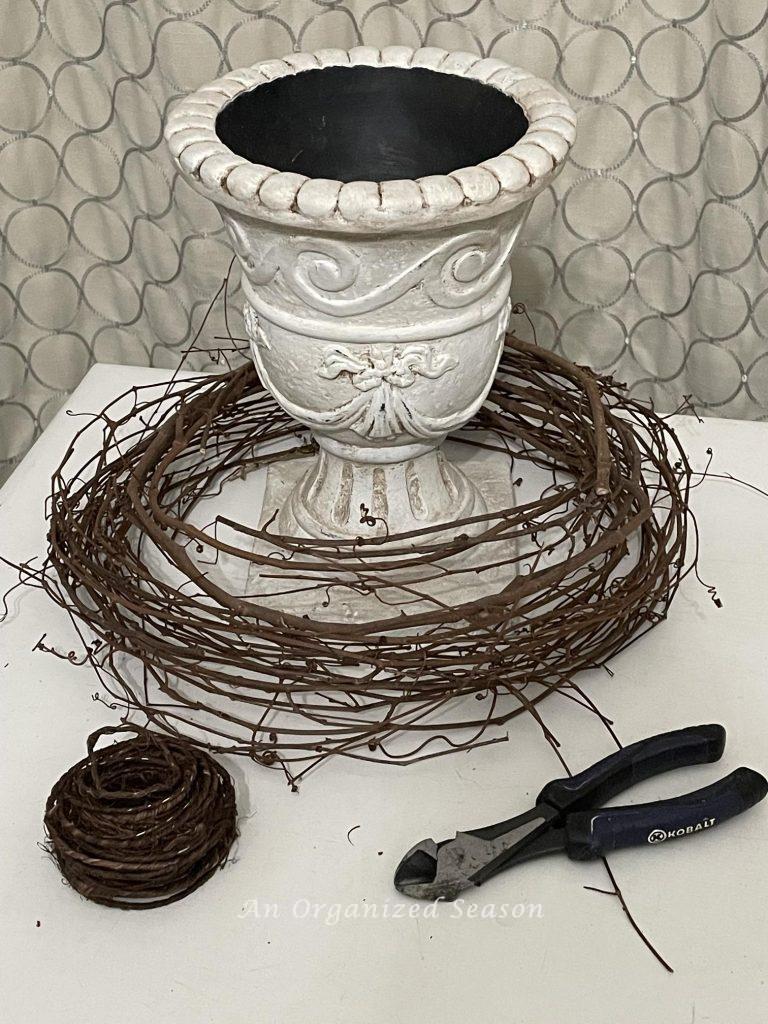Gather an urn, grapevine wire, garpevine wreath, and wire cutters to DIY a topiary frame from a grapevine wreath.