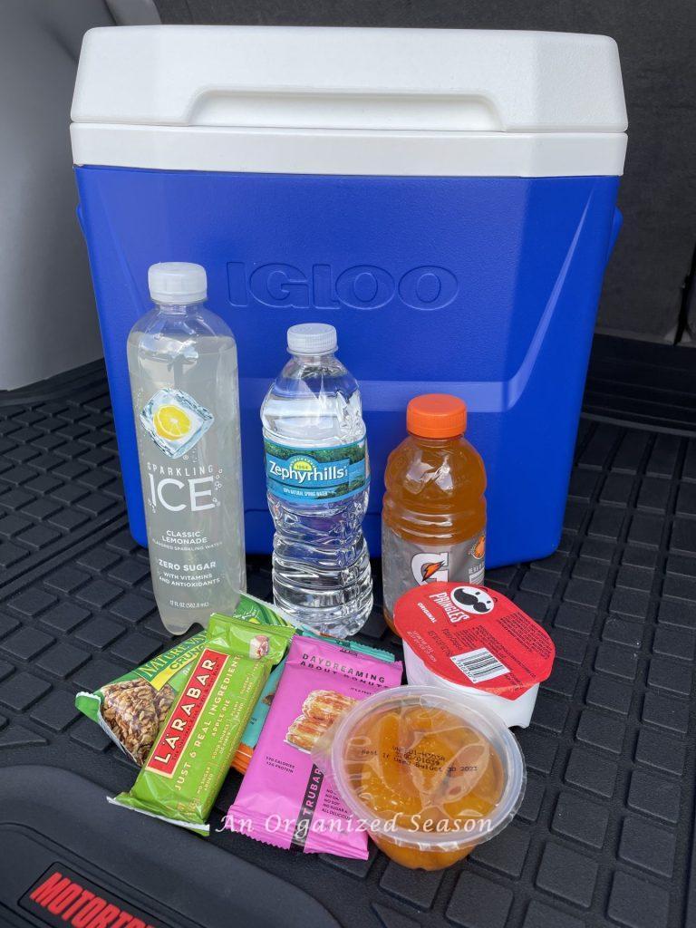 A cooler in the back of a car with drinks and snacks in front of it. 