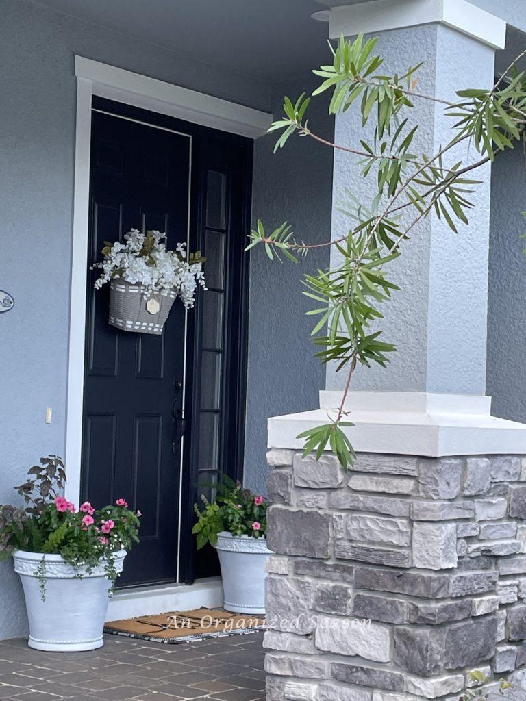 A front porch with a black door that has a floral basket hanging on it. 