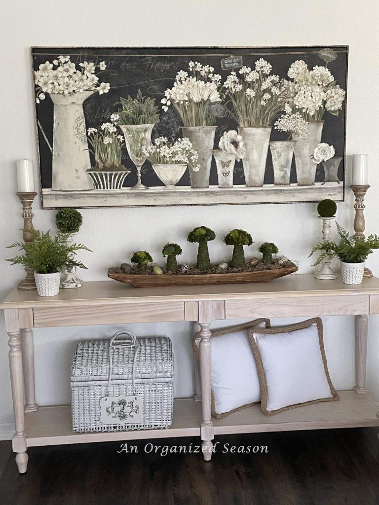 A picture with white flowers hanging over a white-washed console table decorated for Spring. 