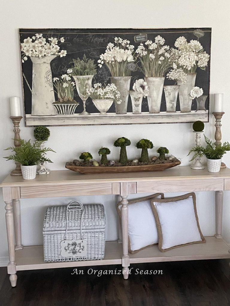 A picture with white flowers hanging over a white-washed console table decorated for Spring.