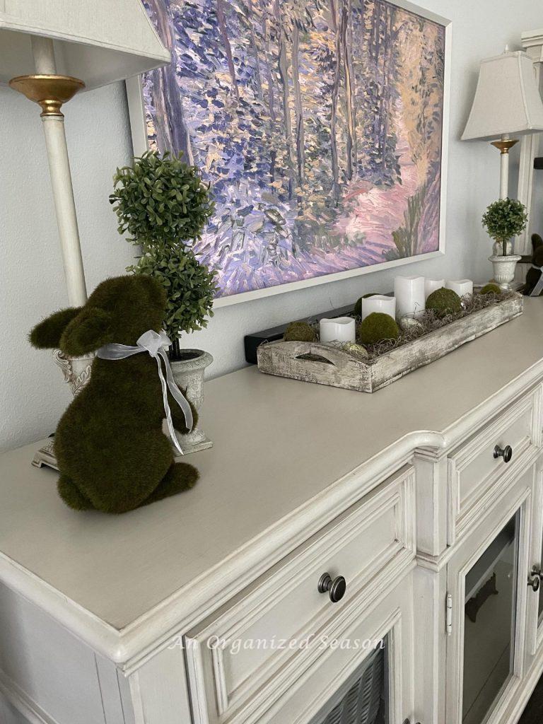 Moss bunnies, topiaries, and a white tray on a TV console table. 