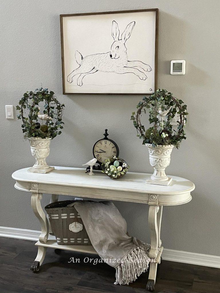 A bunny picture hanging over a foyer table decorated for Spring with two topiaries. 