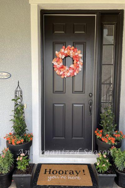 tulip wreath on black door surrounded by orange flowers in pots.