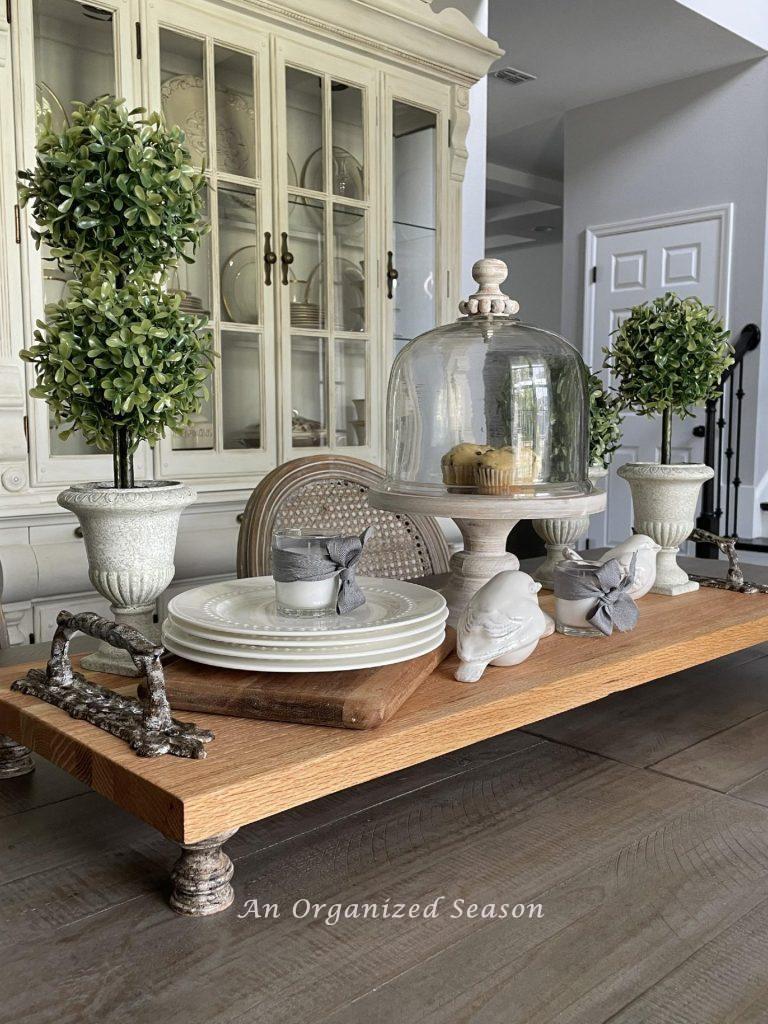A wood tray holding topiaries, muffin stand, plates, and white birds on a dining table. 