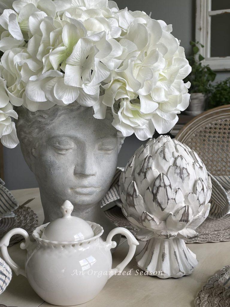 A planter bust filled with white hydrangeas next to an artichoke sculpture and a white sugar bowl. 