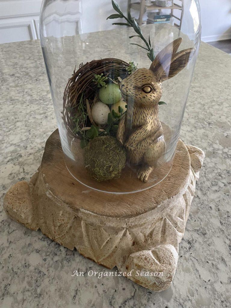 A gold bunny, nest, and moss ball inside a cloche. 
