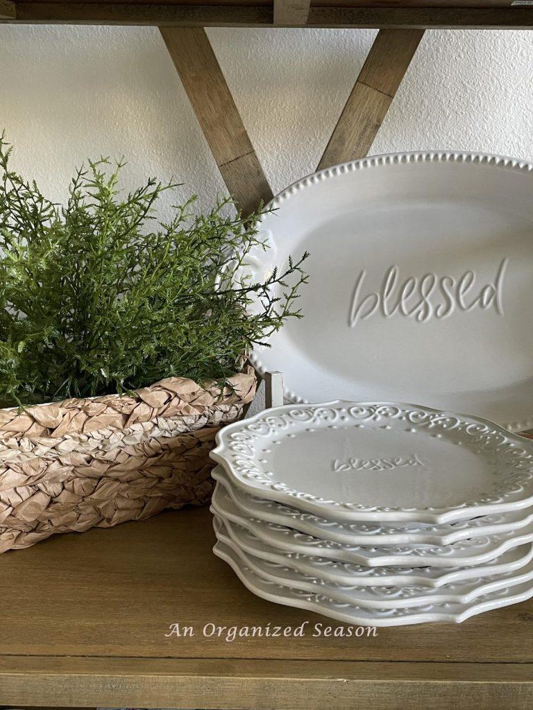 A basket with a faux plant sitting on a shelf with white plates.  