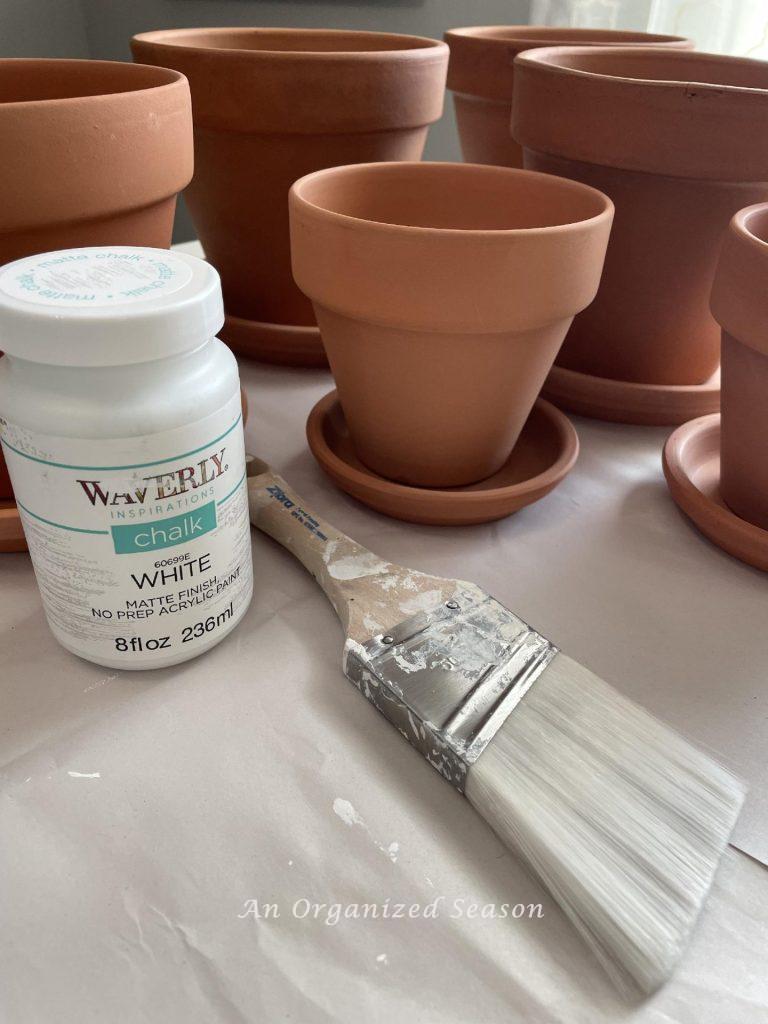 Terra cotta pots, a paintbrush, and white chalk paint are gathered to paint flower pots.