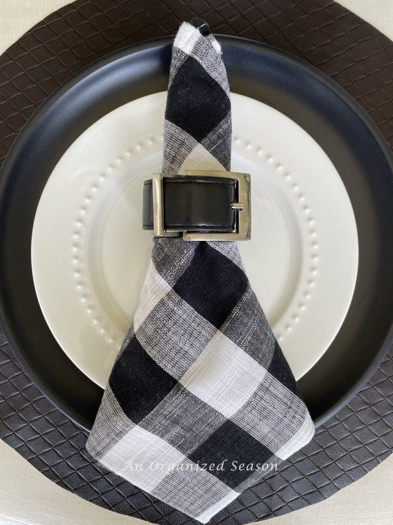 A napkin ring made from a black leather belt around a buffalo check napkin. 