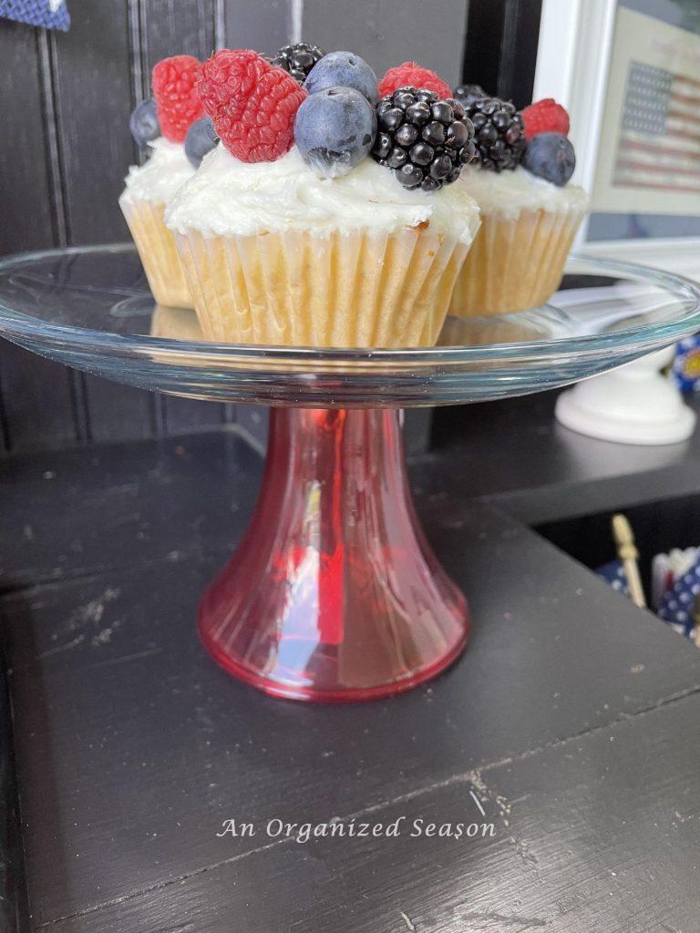 Vanilla cupcakes topped with fruit sitting on a glass cake plate.