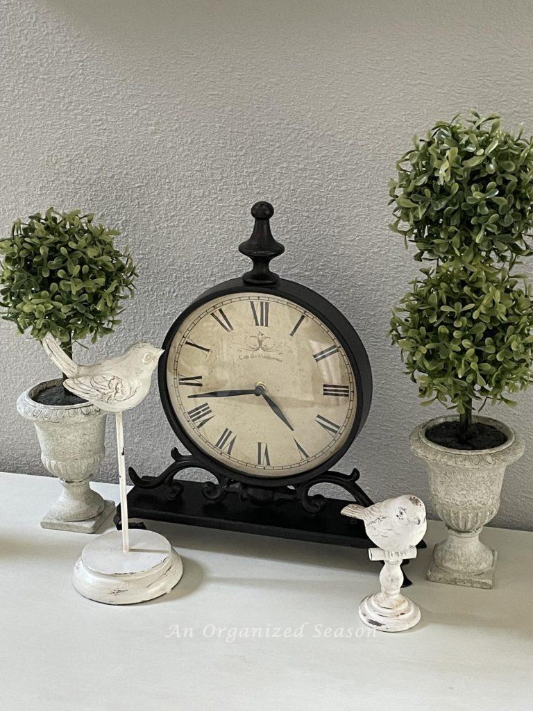 Two small topiaries, a clock and two birds on a pedestal.
