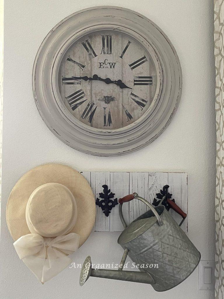 A wall clock above a coat rack that has a sun hat and a watering can hanging from it. 