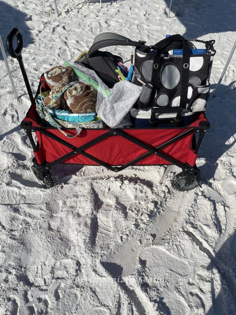 A red collapsible wagon on a beach filled with essential items. 