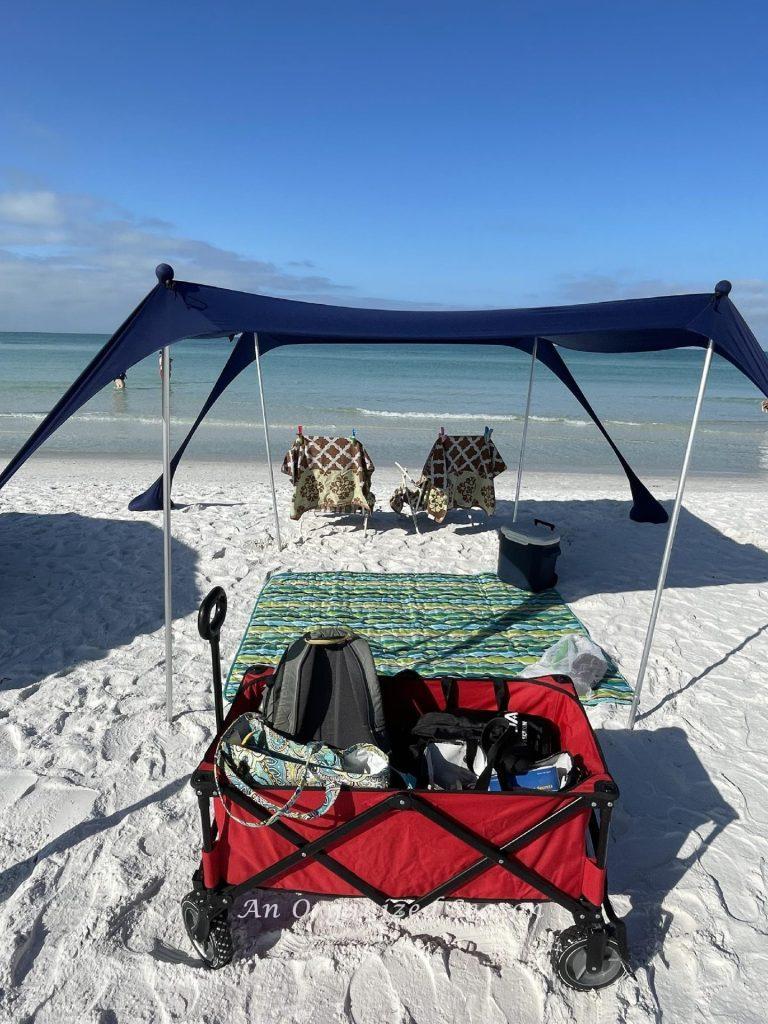 A red wagon, beach blanket, cooler, beach chairs, and a tent set up on the beach. 