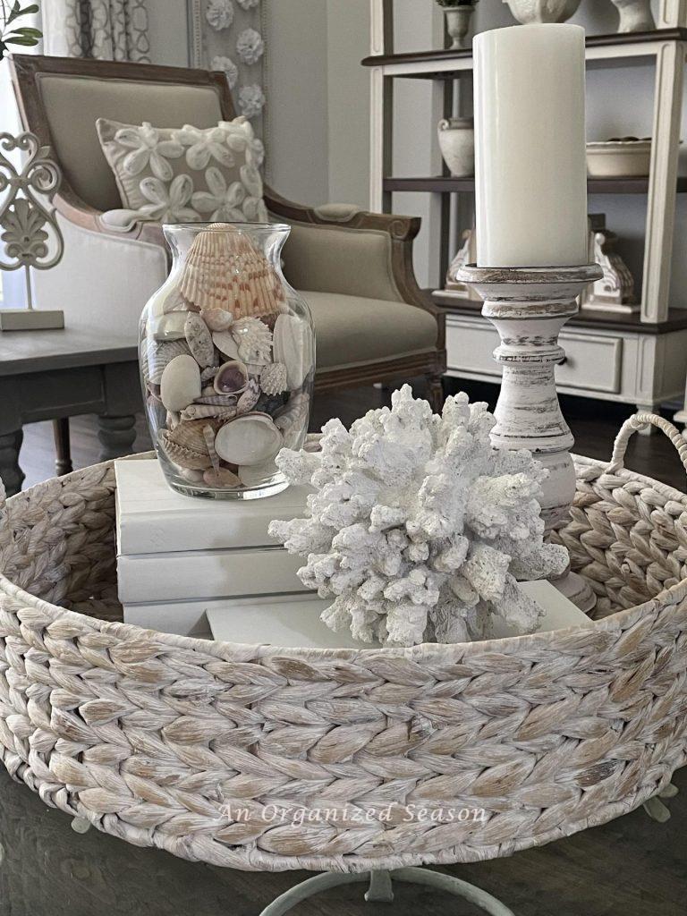 A white-washed basket filled with a candle holder, a glass bowl of seashells and a large piece of coral. 