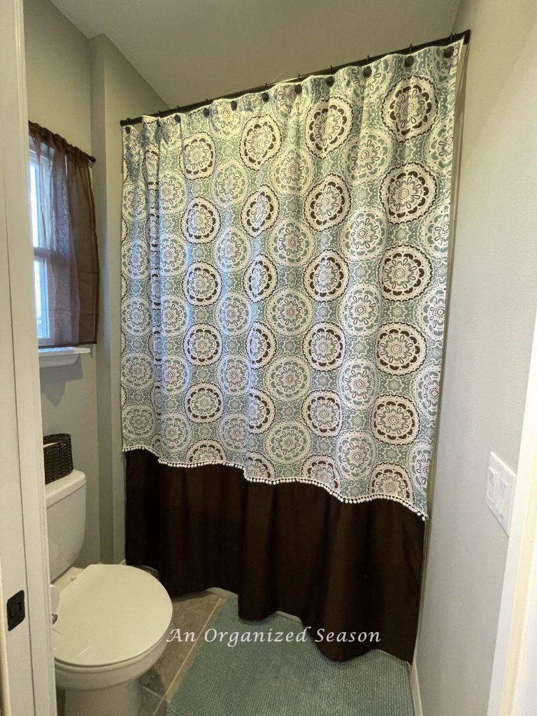 A printed shower curtain lengthened with brown fabric and pompom trim.