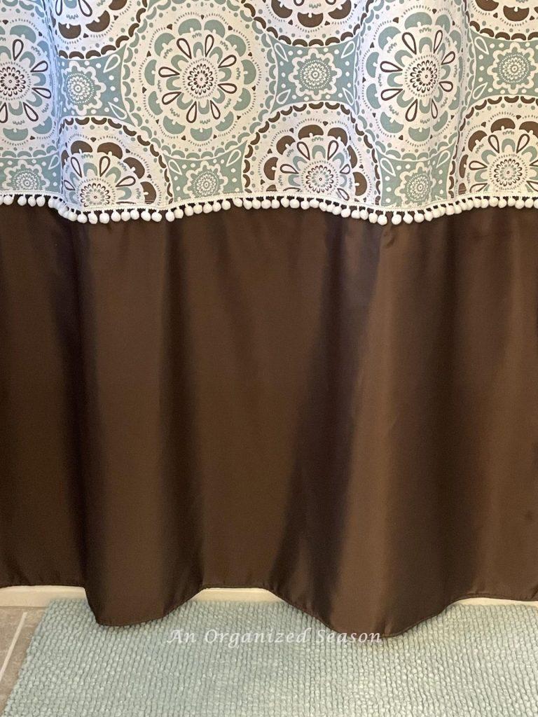 A shower curtain with pompom trim and brown fabric added to the bottom. 