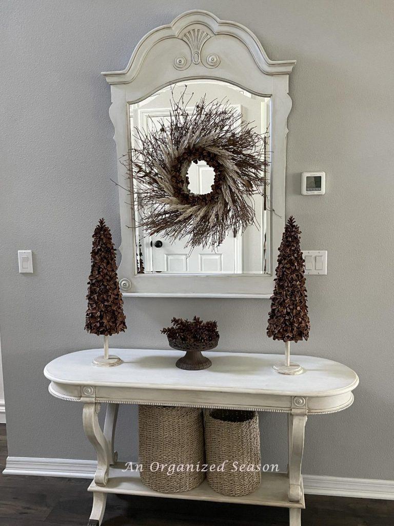 Two pinecones Christmas trees on a foyer table with a pinecone flower wreath hanging on a mirror behind them.