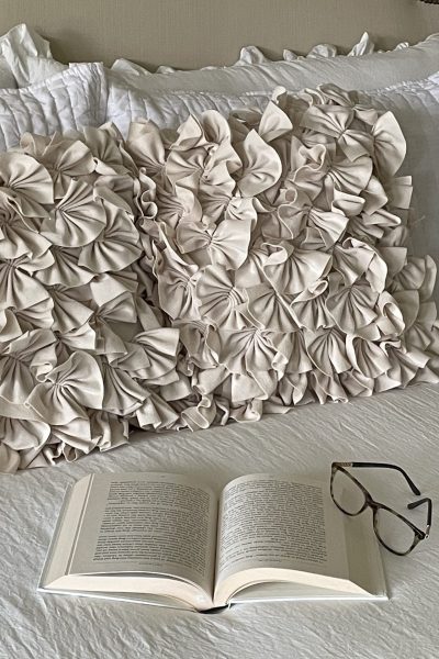 A book and a pair of glasses laying on a bed next to ruffled pillows.