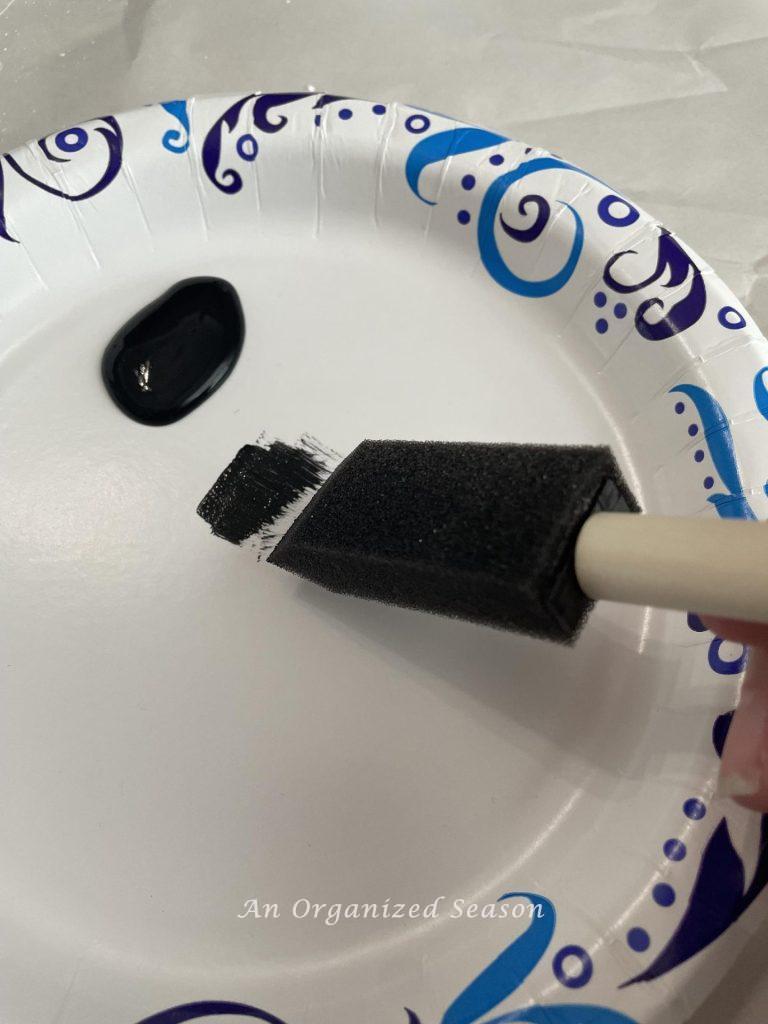 Step two to distress with paint is to add black paint to a paper plate, dip the tip of a sponge in it, and wipe the excess paint off on the plate.