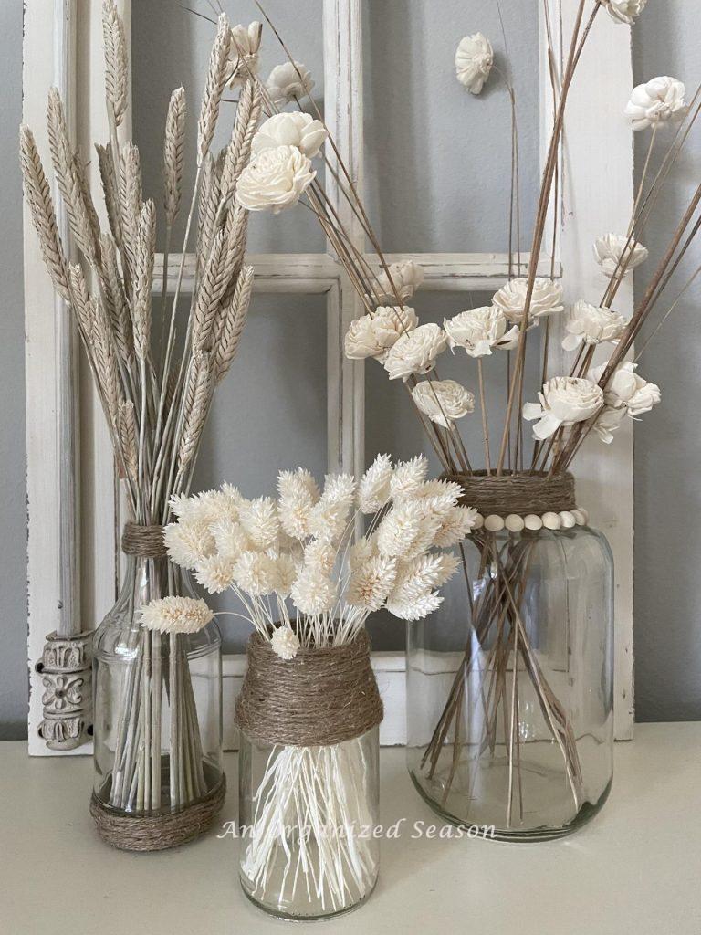 Three glass vases with twine accents holding white dried flowers and wheat. 