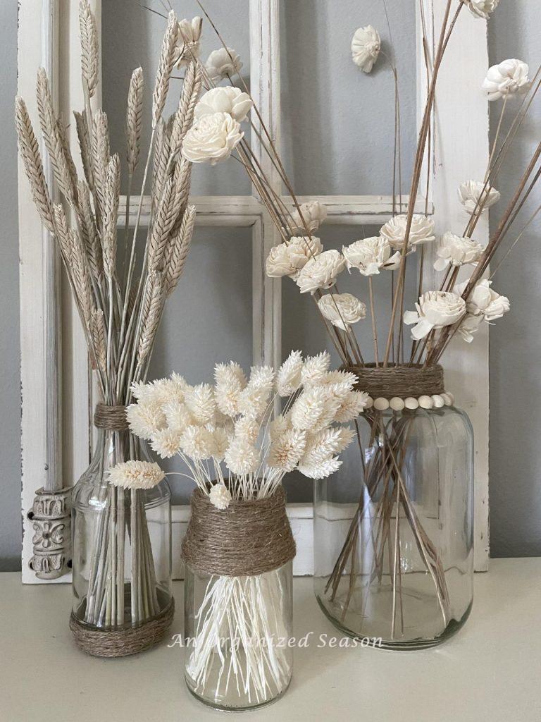Three glass vases with twine accents holding white dried flowers and wheat.