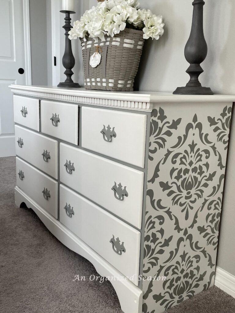 White dresser with gray handles and damask design on the sides. 