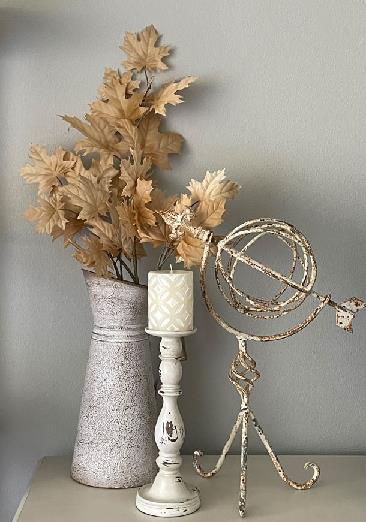 Tan leaves in a white pitcher next to a white candlestick and an iron sculpture.