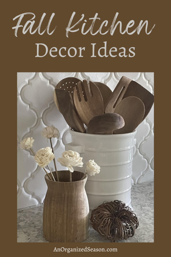 Wooden spoons in a white container next to a wood vase with white faux flowers and a grapevine pumpkin on a kitchen counter. 