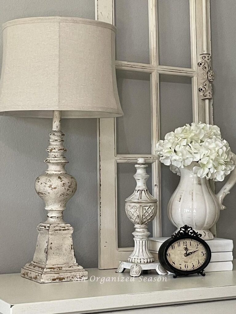 A lamp, clock, floral arrangement, and sculpture on a nightstand. 